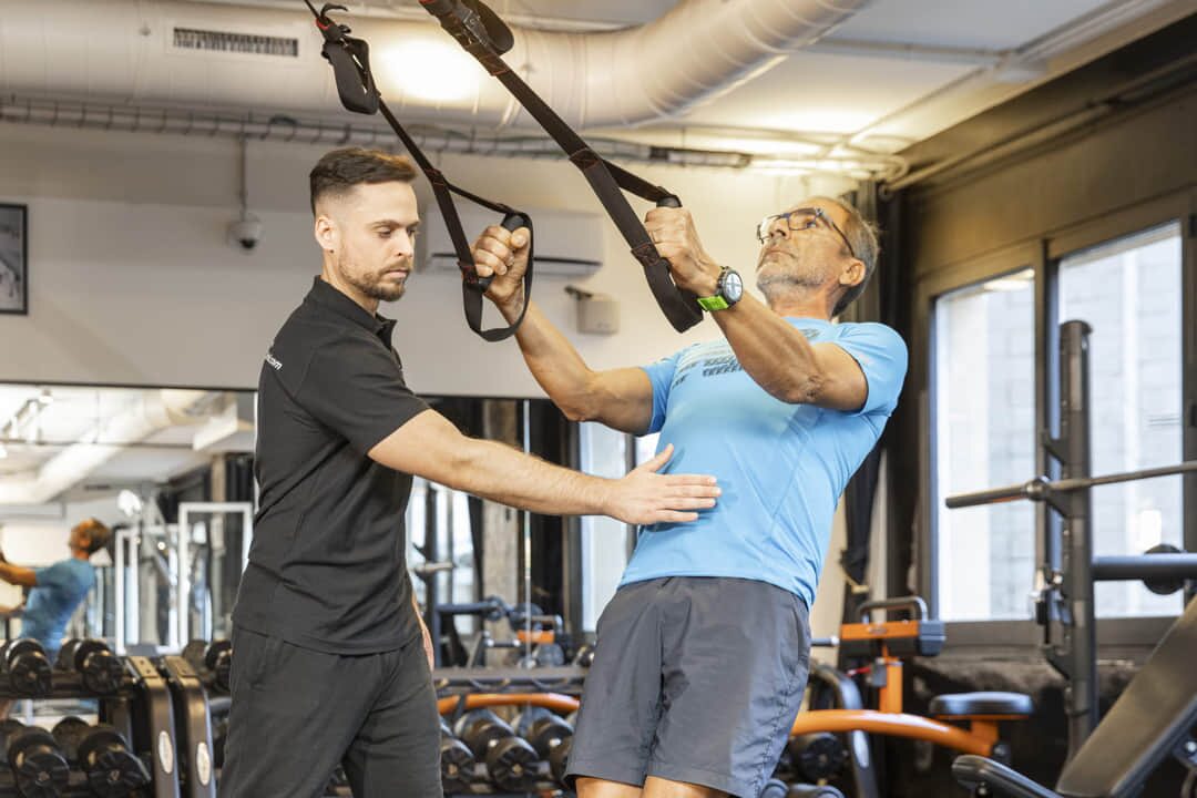 Entrenador personal guía a un cliente senior durante un ejercicio de suspensión TRX en nuestro gimnasio de Barcelona