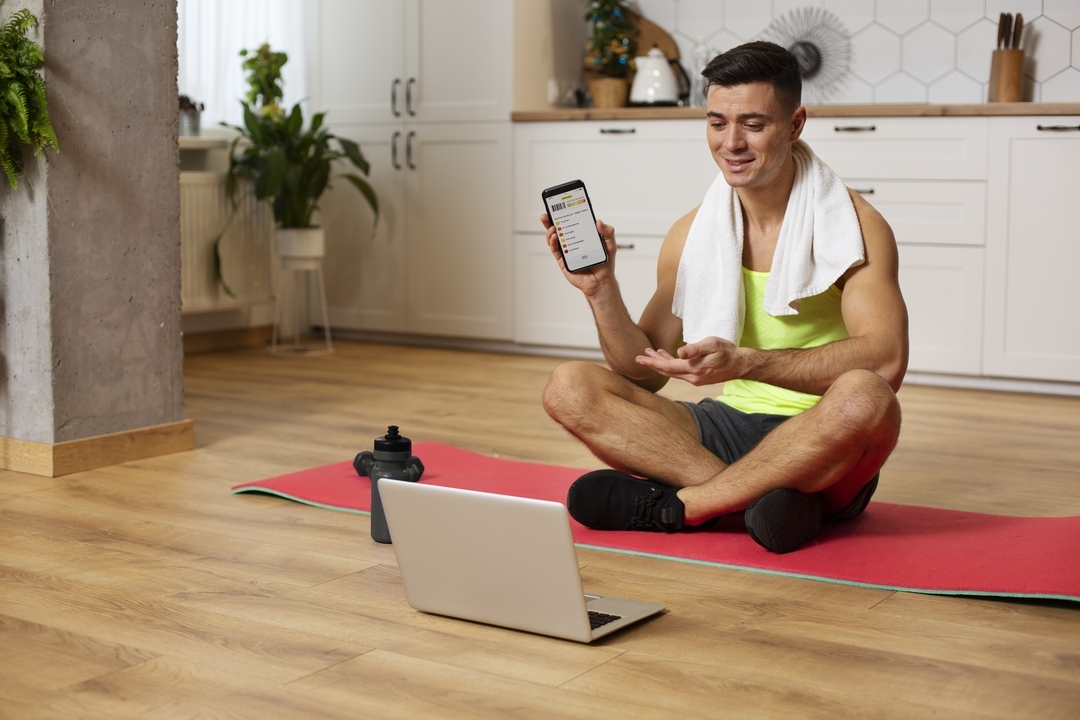 Cliente charla con su entrenador personal a través del portátil sentado en su colchoneta tras la sesión online desde casa en Barcelona, mostrando la rutina en el móvil