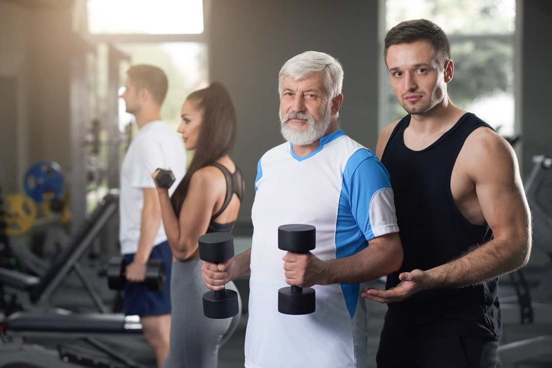 Entrenador acompaña a un adulto mayor mientras trabaja fuerza de brazos con mancuernas en nuestro gimnasio de Barcelona; al fondo, otros alumnos siguen su rutina