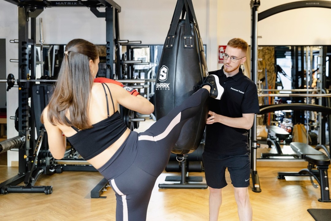 Entrenador sujeta el saco mientras una alumna ejecuta una patada circular de kick boxing, trabajando potencia y coordinación en nuestro gimnasio de Barcelona