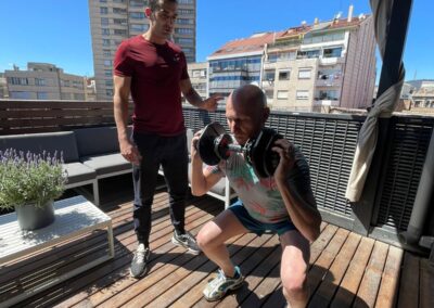 Entrenador guía a un cliente mientras realiza sentadillas con mancuernas en la terraza de un ático en Barcelona, aprovechando el entrenamiento al aire libre