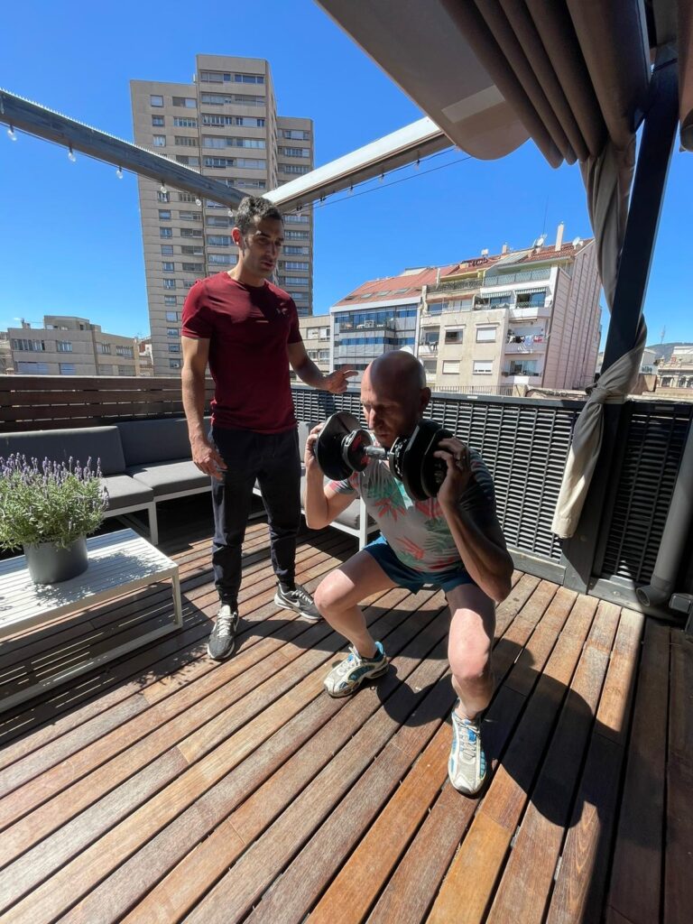 Entrenador guía a un cliente mientras realiza sentadillas con mancuernas en la terraza de un ático en Barcelona, aprovechando el entrenamiento al aire libre